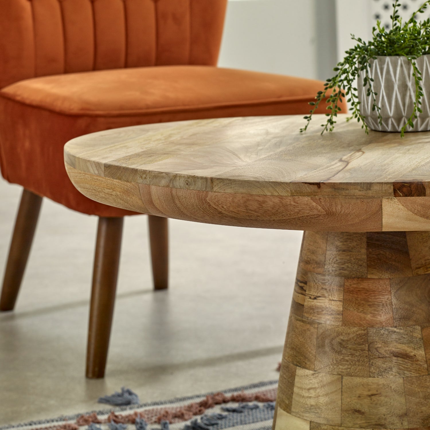 Merlin Mango Wooden Round Coffee Table Mushroom Style Wooden table with a textured surface in a room setting with an orange chair and plant.