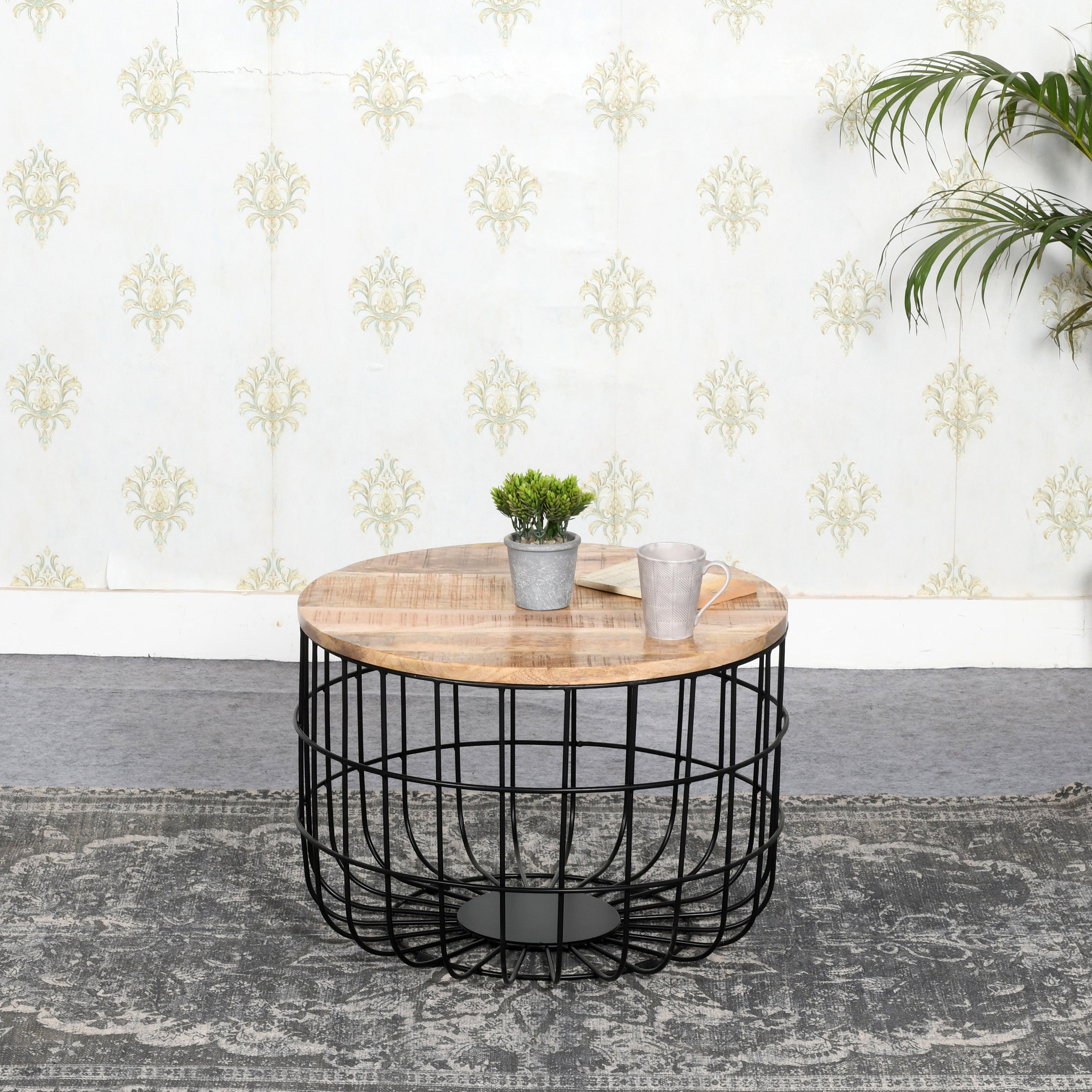 Merlin Mango Wooden Wire Coffee Table Round wooden coffee table with black metal base in a room with patterned wallpaper and rug.
